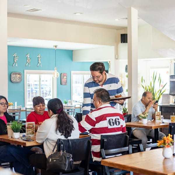A server delivers food to customers seated at wooden tables in a brightly lit Polanquito restaurant.